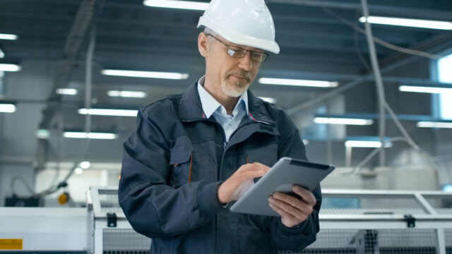 A photograph of a man in glasses, wearing a hardhat and holding a tablet.
