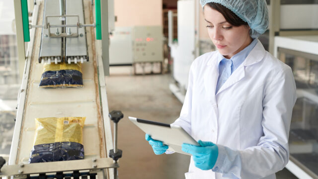 A photograph of a woman in protective gear holding a tablet, performing a quality check on items on the production line.
