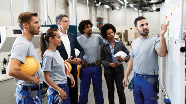Maintenance team talking to a manager and pointing to a production plan on the wall in a factory setting