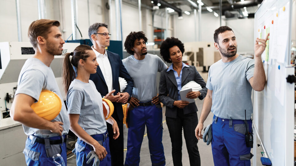 Maintenance team talking to a manager and pointing to a production plan on the wall in a factory setting